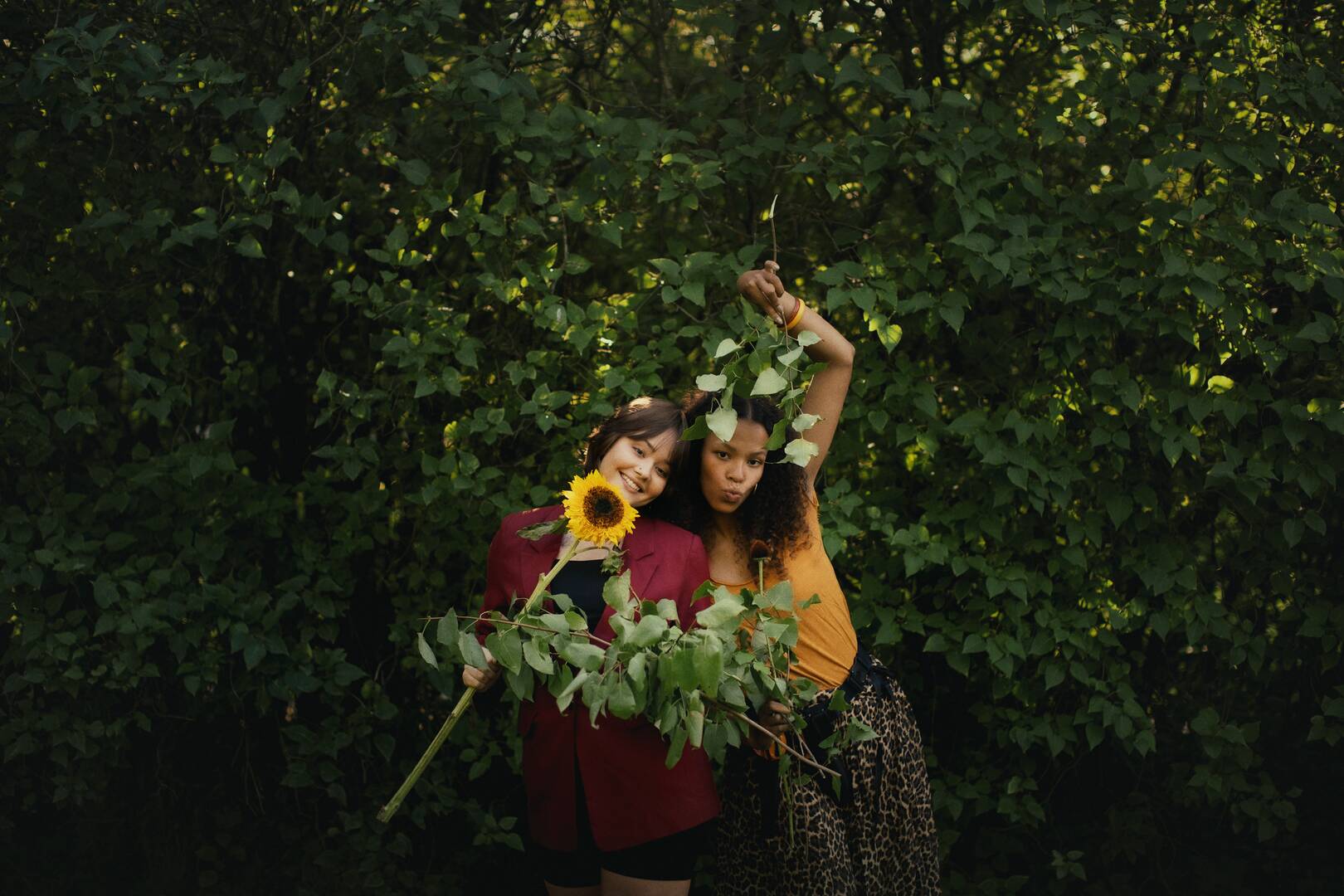 zwei mädchen stehen vor einer grünen hecke und halten eine sonnenblume in der hand