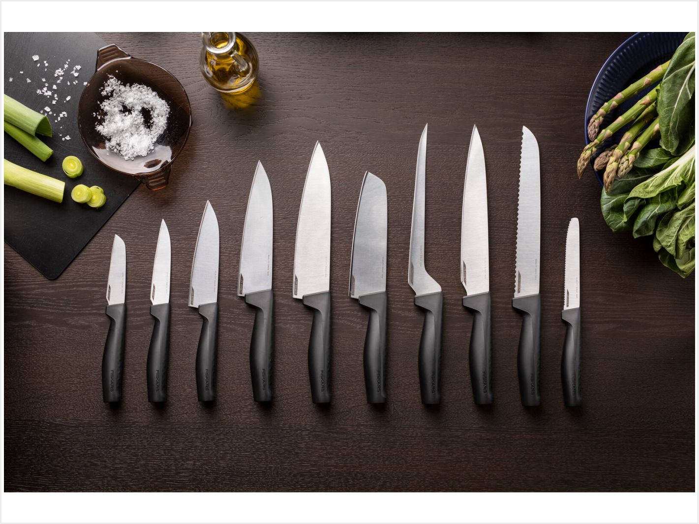 Fiskars Edge Küchenmesser liegen nebeneinander auf einer hölzernen arbeitsfläche. spargel, salz und porree ist mit auf dem bild