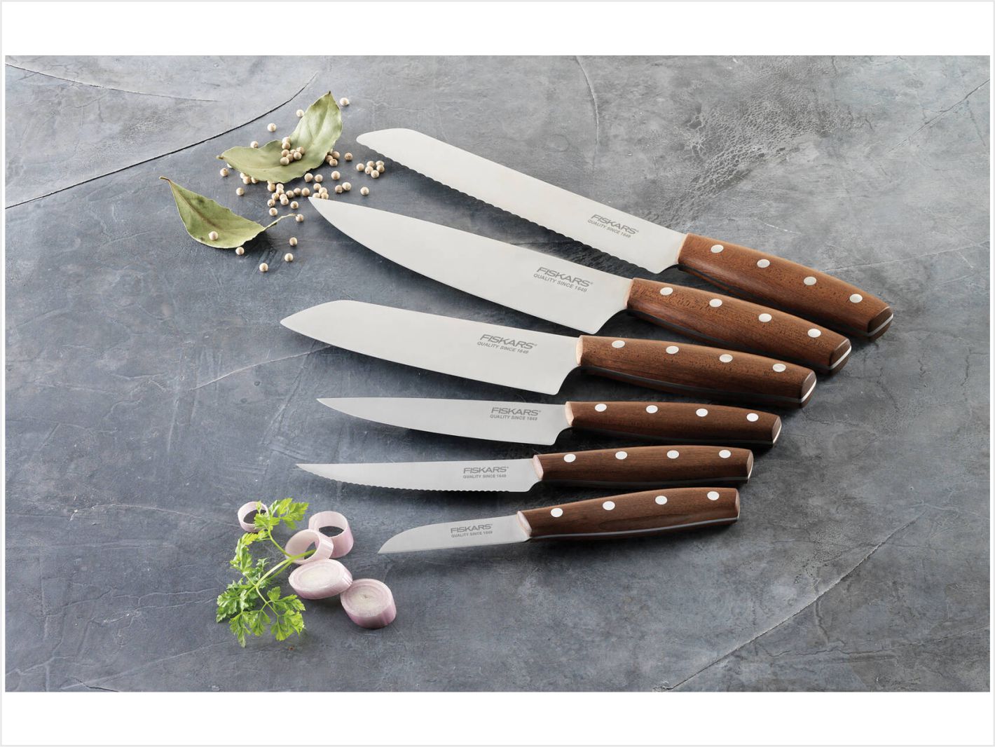 sechs Fiskars Norr Kochmesser auf einer arbeitsfläche. pfefferkörner und etwas schnittlauch liegen verteilt um die messer die individuell graviert werden können.
